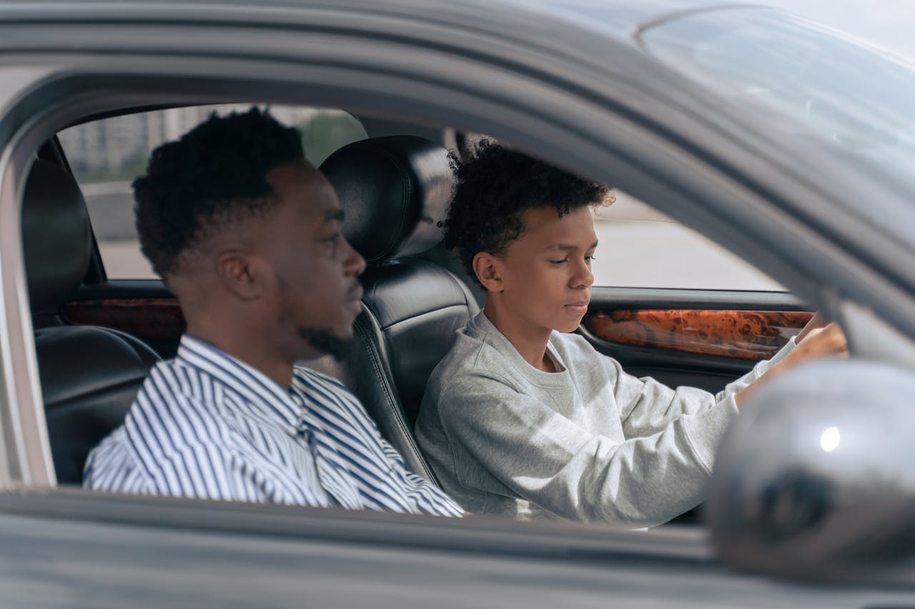 A father sits beside his son who is learning to drive, inside a car.